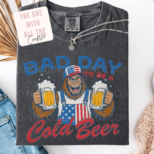 Gray t-shirt with a graphic of bigfoot holding beers and text 'Bad Day to be a Cold Beer' on a white background.