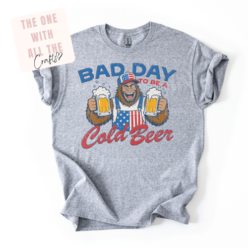 Gray t-shirt with a graphic of bigfoot holding beers and text 'Bad Day to be a Cold Beer' on a white background.