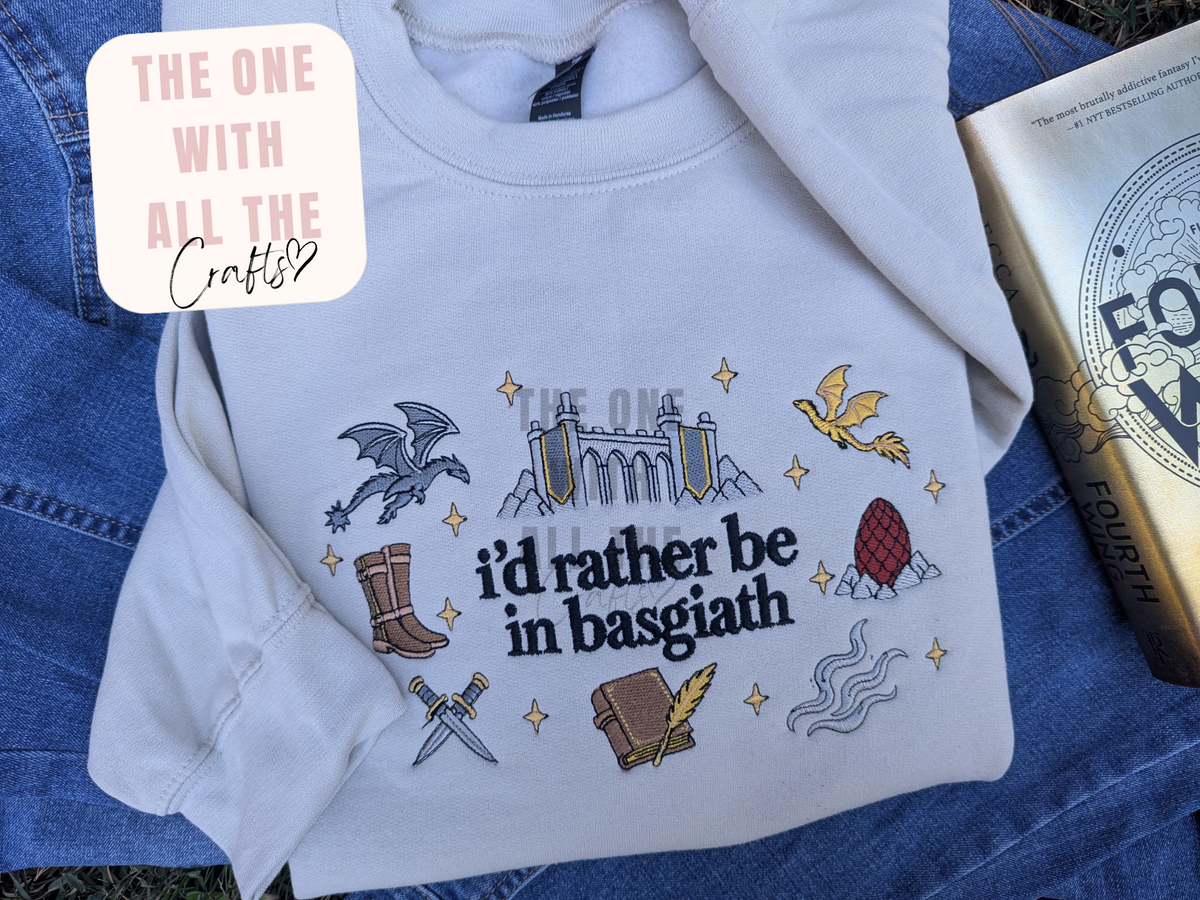 A light gray long-sleeve T-shirt laid flat on blue denim, featuring embroidered or printed fantasy-themed illustrations and the text 'i'd rather be in basgiath.' A white badge in the top left reads 'THE ONE WITH ALL THE Craft&Co,' and a book is partially visible on the right.