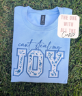 A folded light blue t-shirt laid flat on green grass, featuring the phrase 'can't steal my JOY' in blue and white floral block letters. A brand tag for Gildan is visible inside the collar, and a decorative sticker reading 'THE ONE WITH ALL THE Crafts' is placed on the upper right.