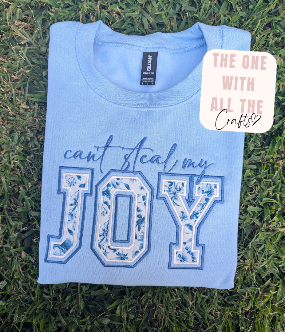 A folded light blue t-shirt laid flat on green grass, featuring the phrase 'can't steal my JOY' in blue and white floral block letters. A brand tag for Gildan is visible inside the collar, and a decorative sticker reading 'THE ONE WITH ALL THE Crafts' is placed on the upper right.