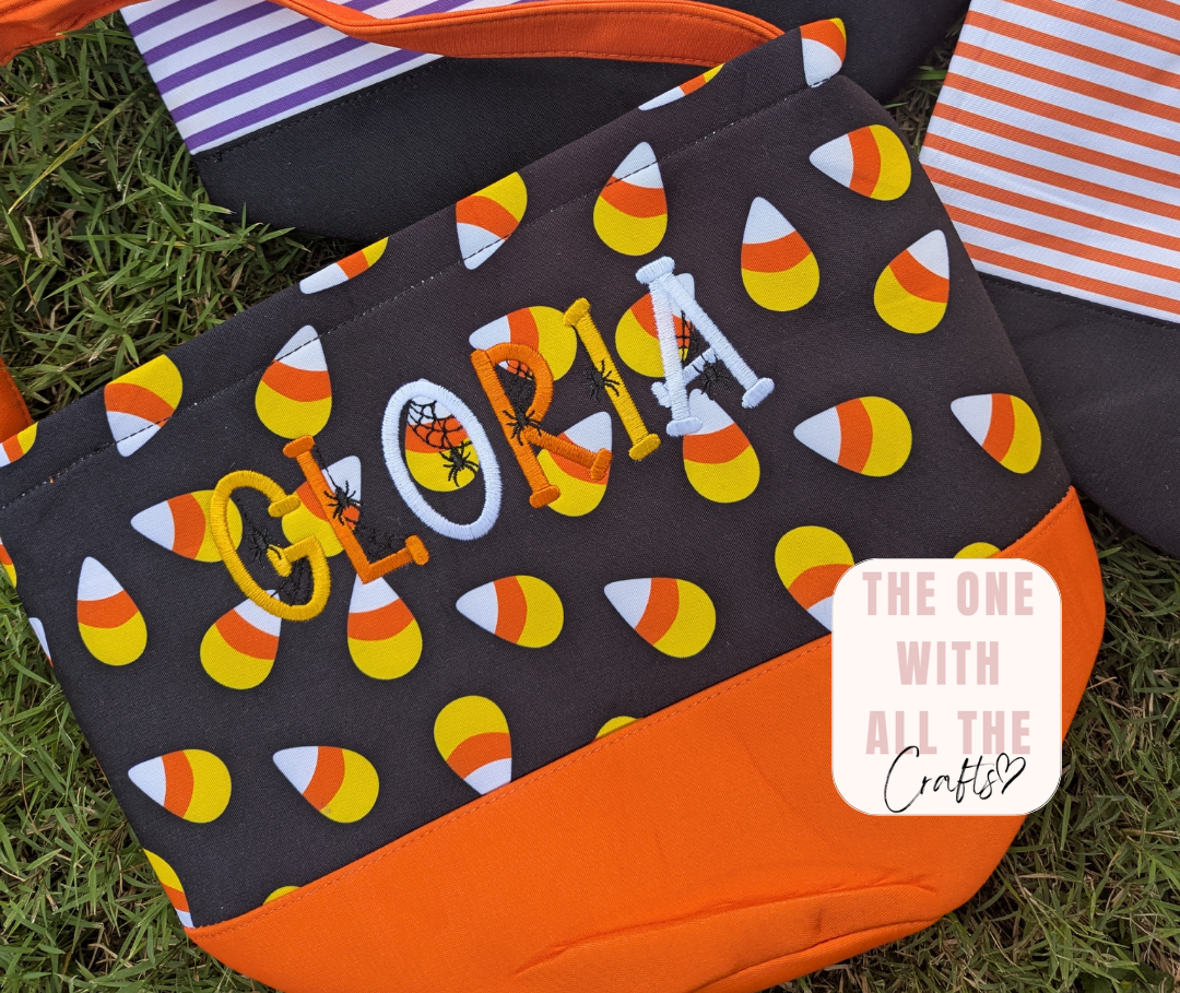 A black fabric tote bag with an orange bottom panel, decorated with a candy corn pattern and the name 'GLORIA' in embroidered orange, white, and black letters. The bag is placed on green grass next to other patterned bags, with a 'The One With All The Crafts' logo sticker in the lower right corner.