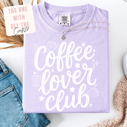 Lavender t-shirt with 'Coffee Lover Club' text on a white surface with denim jeans and decorative items.