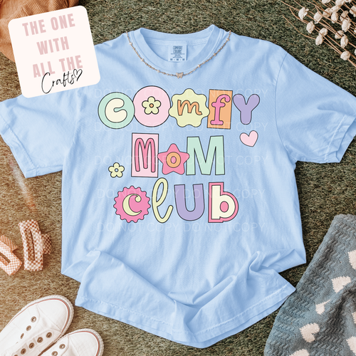 Light blue t-shirt with 'Comfy Mom Club' text on a textured surface with accessories.