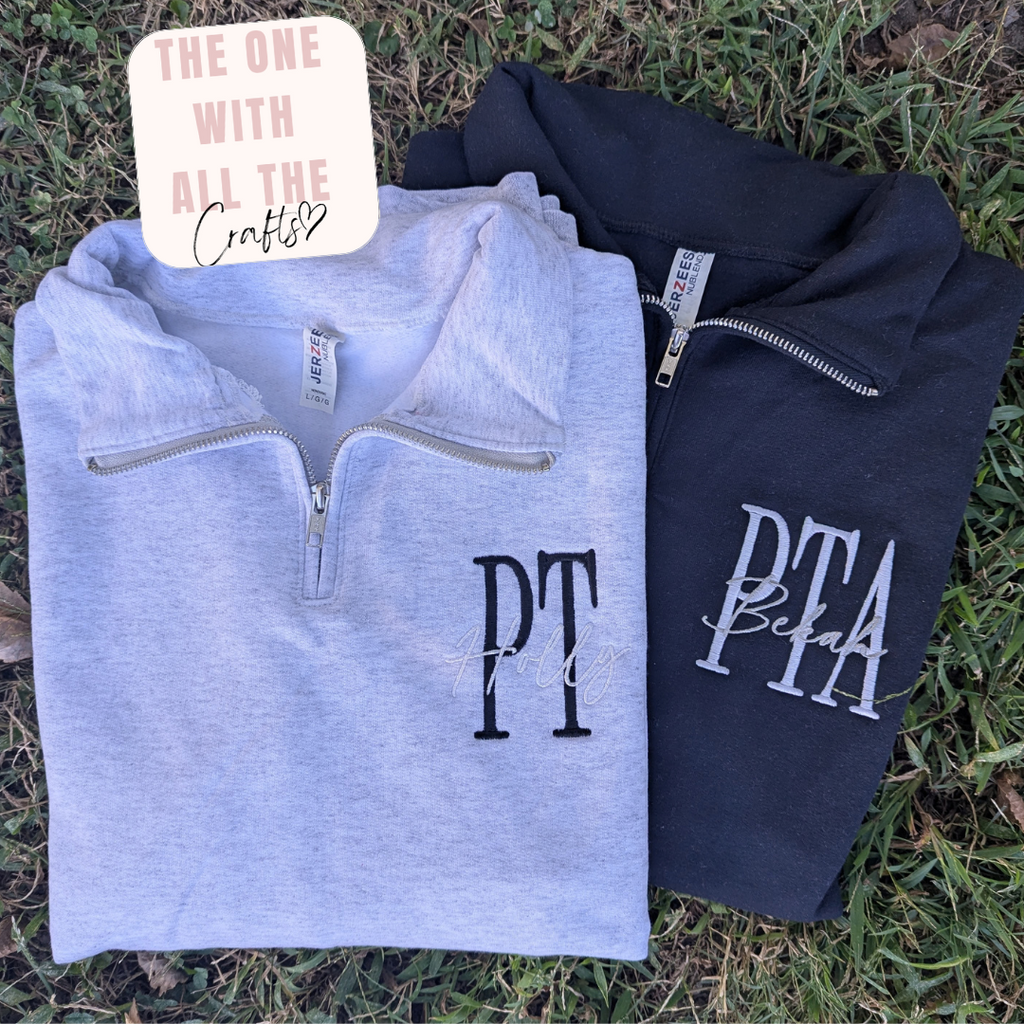 Two folded quarter-zip sweatshirts laid flat on grass: a light heather gray one with black 'PT' lettering and a navy one with white 'PTA' lettering. A cream badge in the top left reads 'THE ONE WITH ALL THE Crafts'.