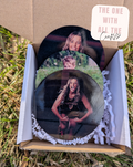 Three circular photo buttons with a person's image on them, placed in an open cardboard box outdoors.