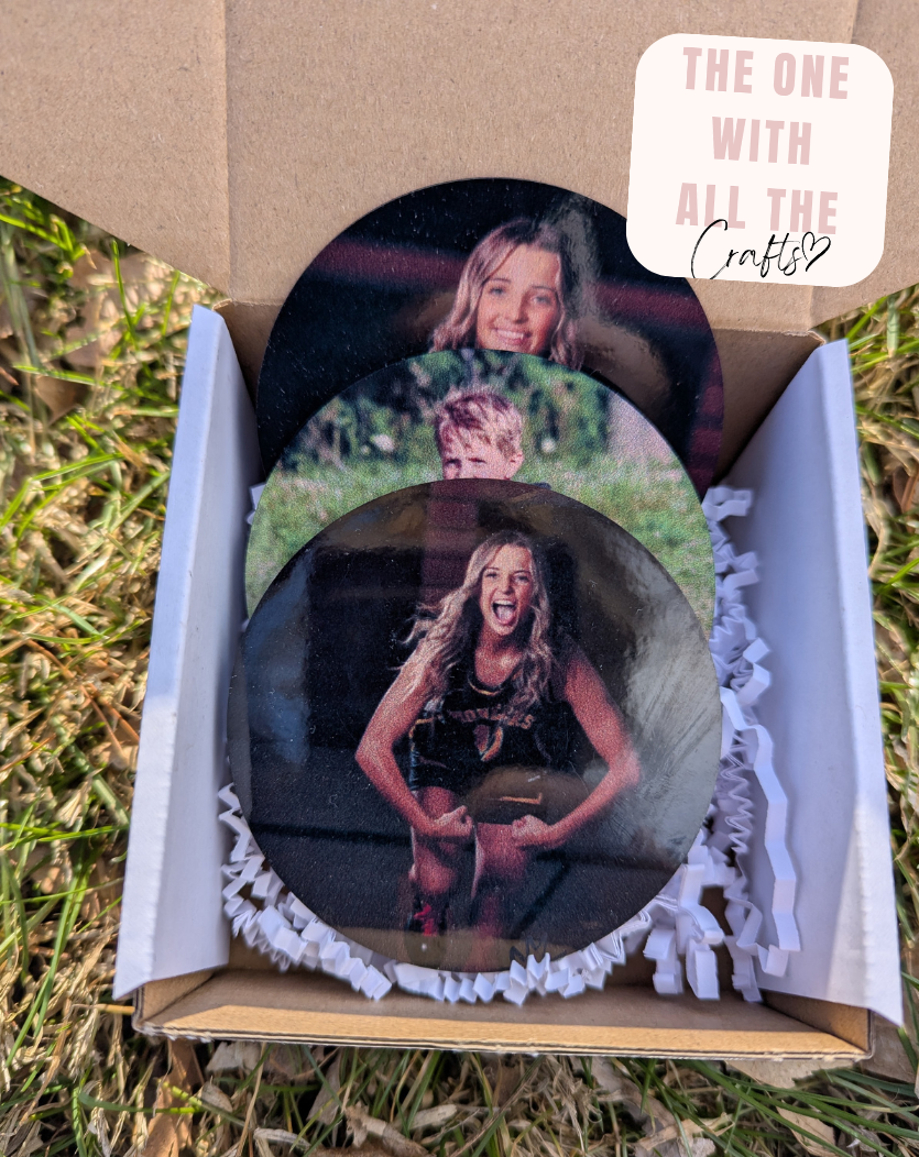 Three circular photo buttons with a person's image on them, placed in an open cardboard box outdoors.