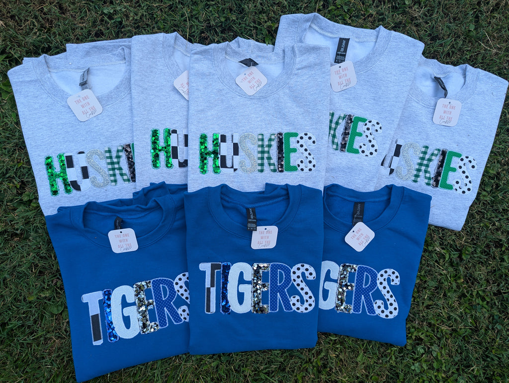 Six short-sleeve graphic T-shirts laid flat on grass, alternating between light heather gray and royal blue, each printed with large 'HUSKIES' or 'TIGERS' lettering in mixed animal-print and camouflage patterns.