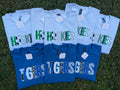 Six short-sleeve graphic T-shirts laid flat on grass, alternating between light heather gray and royal blue, each printed with large 'HUSKIES' or 'TIGERS' lettering in mixed animal-print and camouflage patterns.