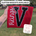 A hand holds a red custom fleece blanket featuring a large black sequined letter 'V' and the word 'vikings' in white script. The background is a grassy outdoor backyard with trees, and there are promotional text overlays at the top and bottom.