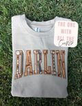 A folded light brown sweatshirt laid on green grass, featuring the word 'DARLIN' across the front in large brown western-style letters with decorative geometric and star details. A pink sticker reading 'THE ONE WITH ALL THE Crafts' is placed on the right side of the sweatshirt.