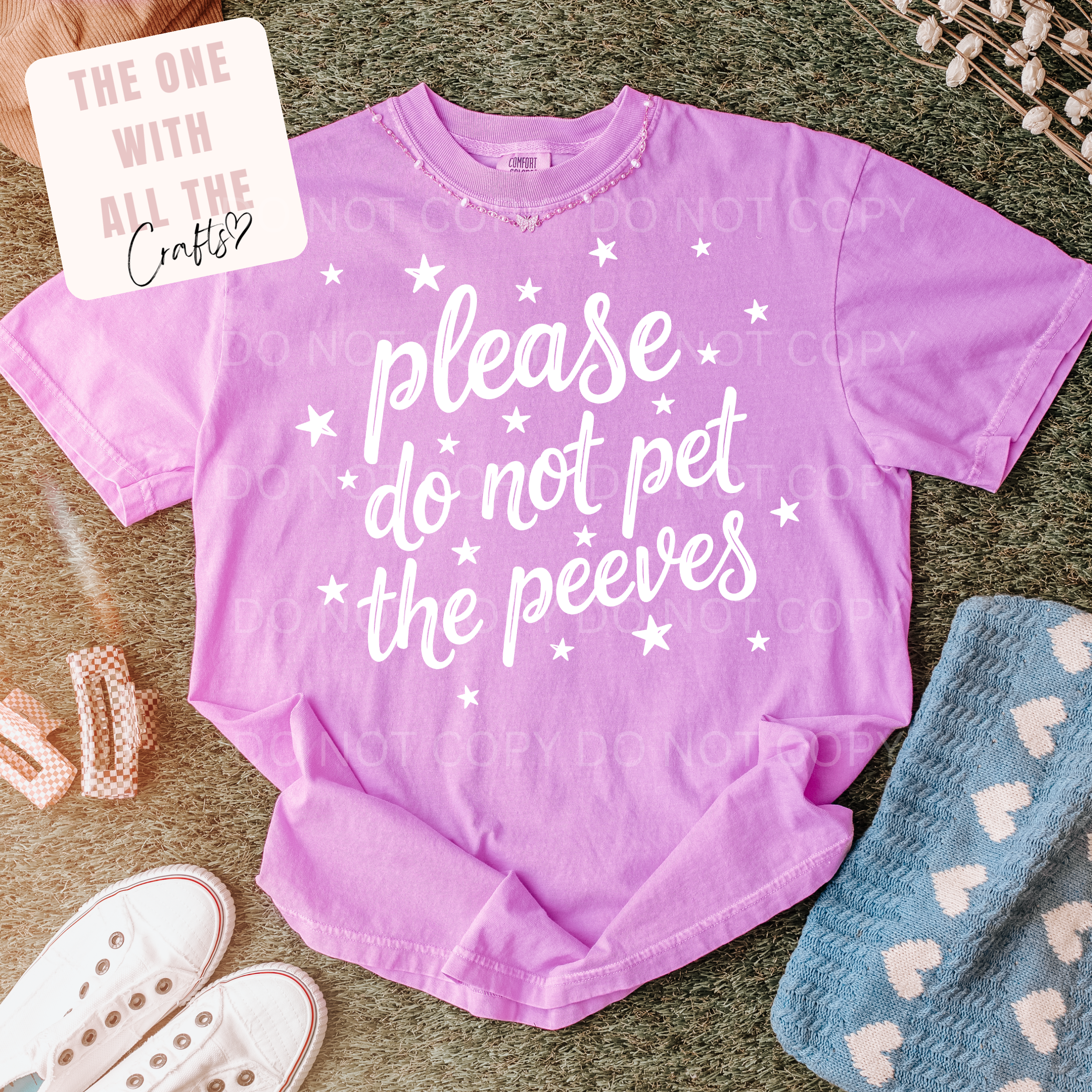 Purple t-shirt with text 'please do not pet the peeves' on a textured surface with accessories.