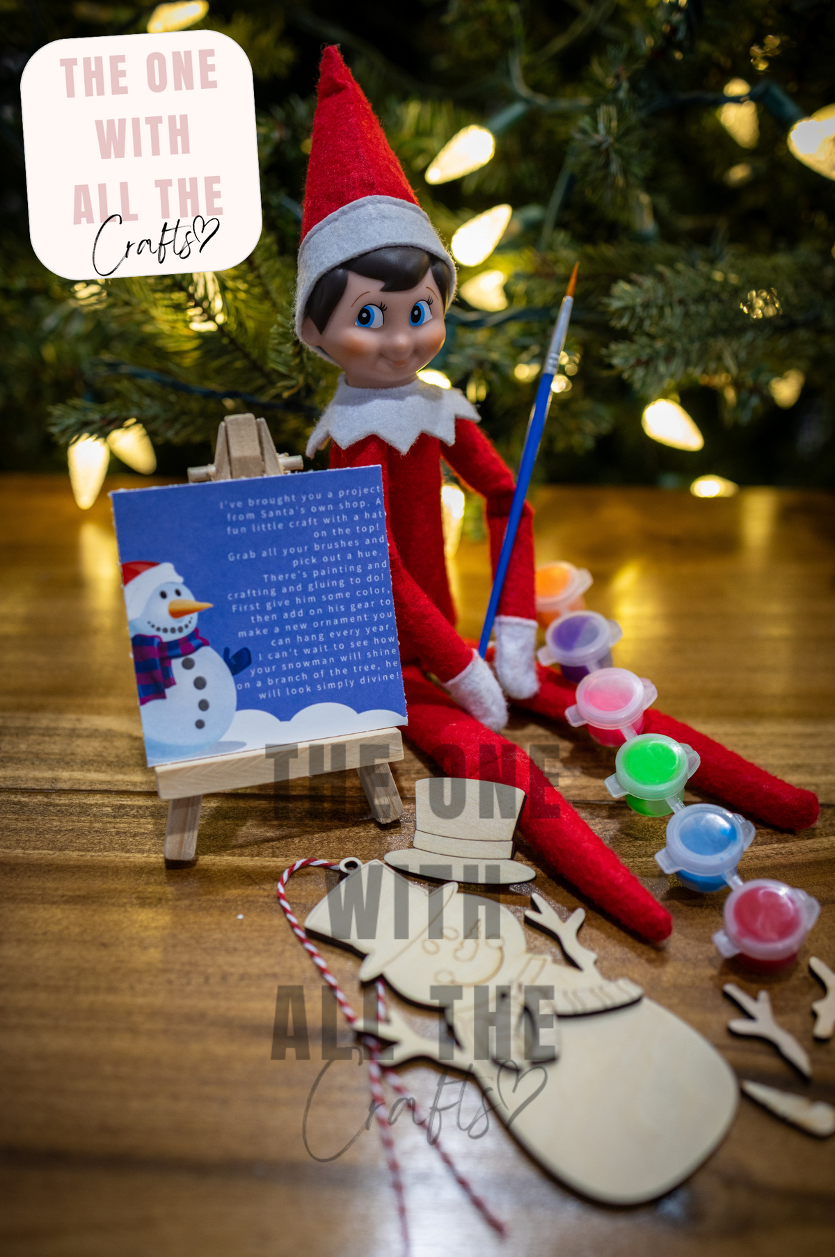 A Christmas-themed craft scene with an elf doll sitting at a small easel holding a blue paintbrush, surrounded by wooden cutouts, paint pots, and a sign that reads 'The One with All the Crafts.' A blurred Christmas tree with warm lights is in the background.