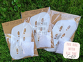 Three packaged 'Elf Footprint Kit' items arranged on green grass, including white footprint-shaped masks or wraps in clear plastic with kraft paper backing and a small white textured piece. A sticker reading 'THE ONE WITH ALL THE CRAFTS' is also shown.