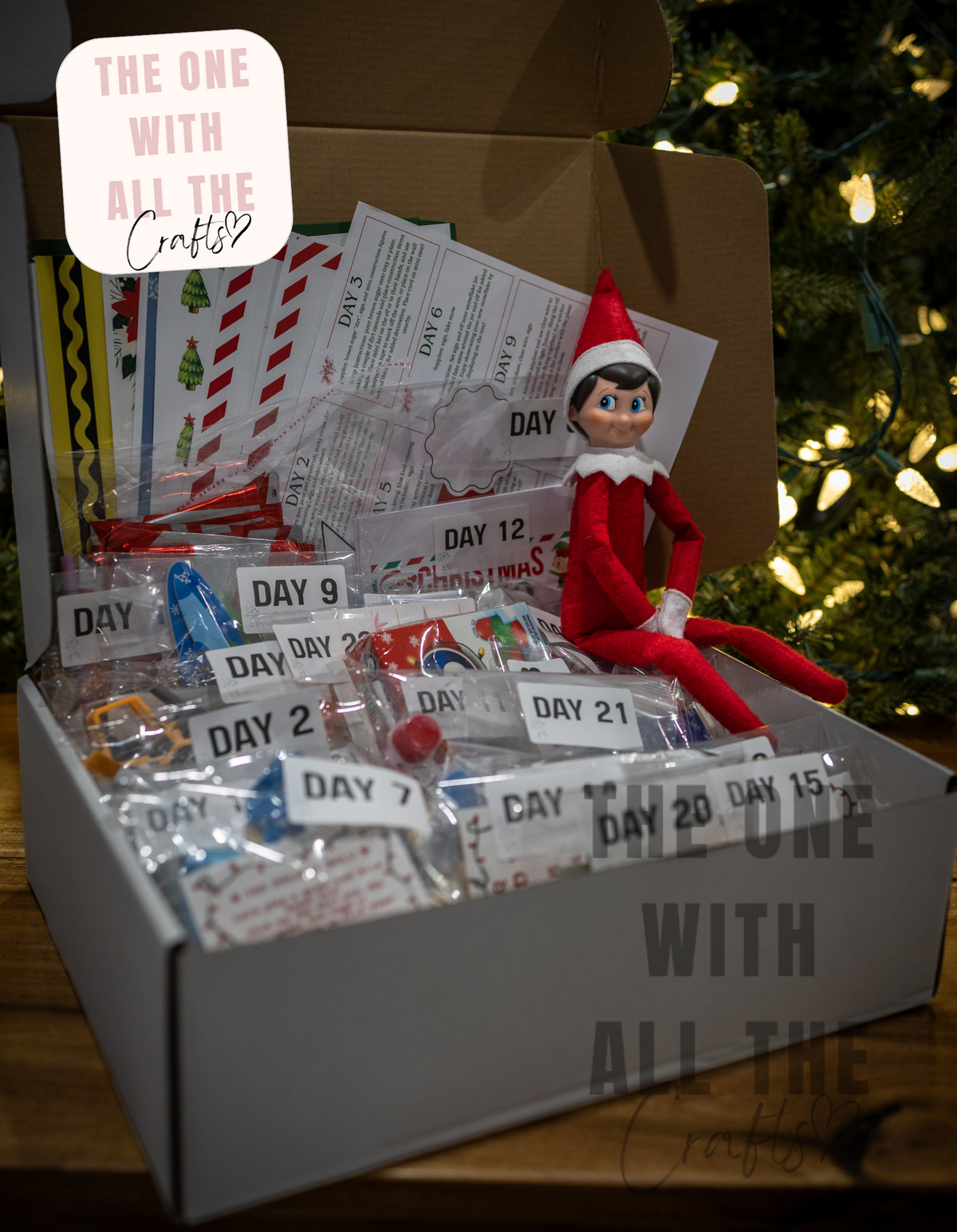 An Elf on the Shelf themed Christmas gift box filled with daily numbered craft challenge packets, small wrapped treats, and a red Elf on the Shelf figure sitting on the edge of the box. The box is set on a wooden surface in front of a blurred Christmas tree with warm lights.