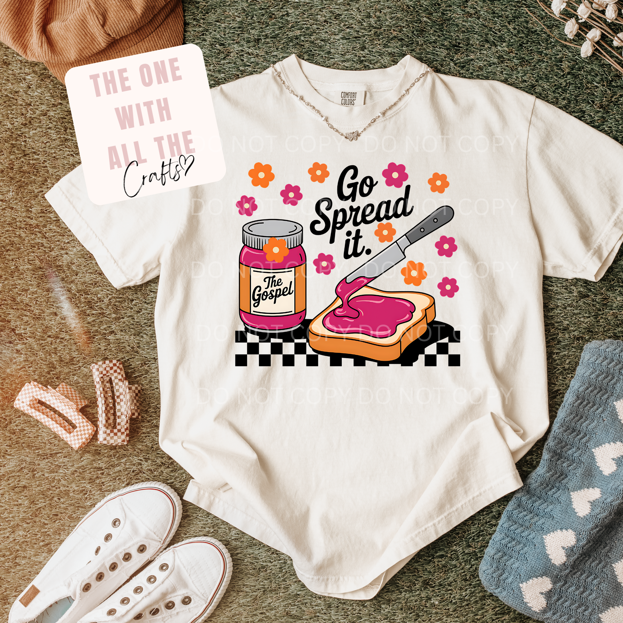 White t-shirt with a graphic design of a jar of jelly and a knife spreading it on toast, surrounded by accessories on a textured surface.
