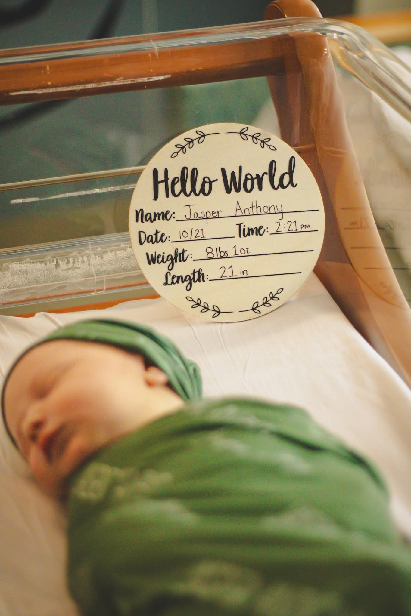 Newborn baby swaddled in green blanket with 'Hello World' milestone card