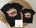 Two black t-shirts with colorful graphics and text on a textured surface.