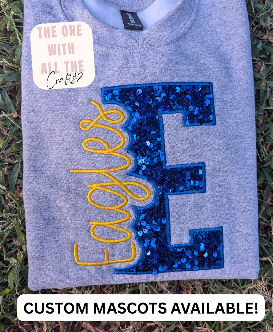 A folded heather gray sweatshirt laid on grass, featuring a large sequined blue letter 'E' with yellow embroidered script text reading 'Eagles' across it. A cream sticker on the shirt reads 'THE ONE WITH ALL THE Crafty', and a white banner at the bottom says 'CUSTOM MASCOTS AVAILABLE!'.