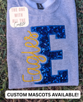 A folded heather gray sweatshirt laid on grass, featuring a large sequined blue letter 'E' with yellow embroidered script text reading 'Eagles' across it. A cream sticker on the shirt reads 'THE ONE WITH ALL THE Crafty', and a white banner at the bottom says 'CUSTOM MASCOTS AVAILABLE!'.
