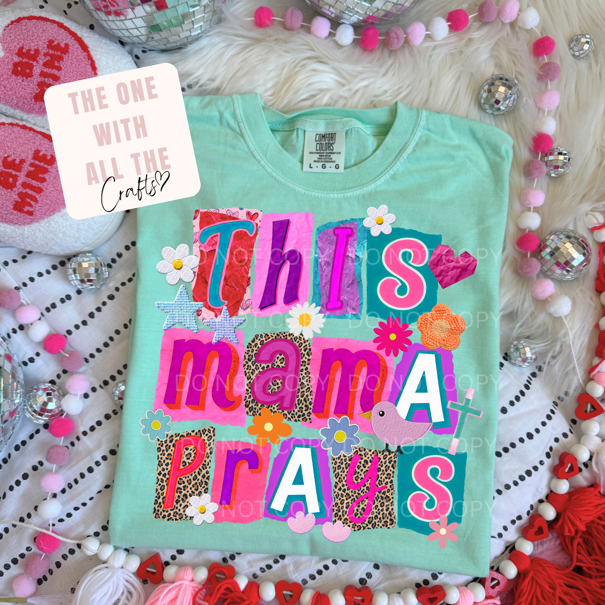 Light blue t-shirt with colorful text 'This mama prays' on a decorative background with hearts and beads.
