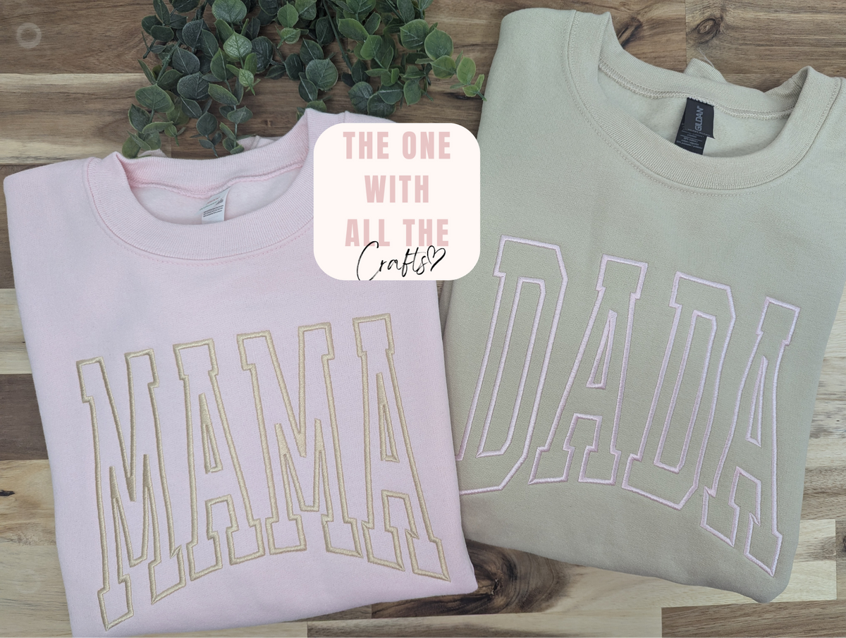 Two folded crewneck sweatshirts displayed on a wooden surface with a green plant in the background. The left sweatshirt is light pink with 'MAMA' outlined in gold.