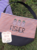 A striped fabric tote bag laid on grass, with orange and white horizontal stripes, a black bottom panel, a black shoulder strap, and embroidered ghosts above the name “FISHER” in black. A purple-and-white striped item is partially visible behind it, and a white sticker reads “THE ONE WITH ALL THE Crafty.