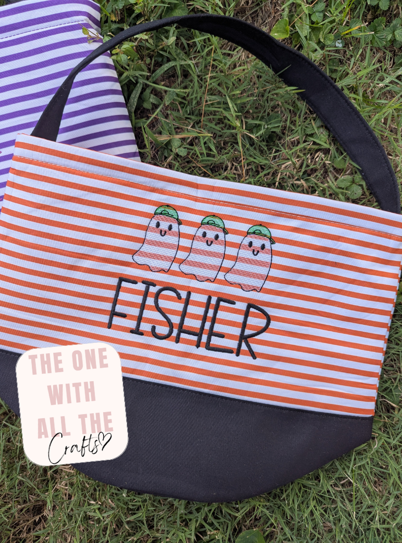 A striped fabric tote bag laid on grass, with orange and white horizontal stripes, a black bottom panel, a black shoulder strap, and embroidered ghosts above the name “FISHER” in black. A purple-and-white striped item is partially visible behind it, and a white sticker reads “THE ONE WITH ALL THE Crafty.
