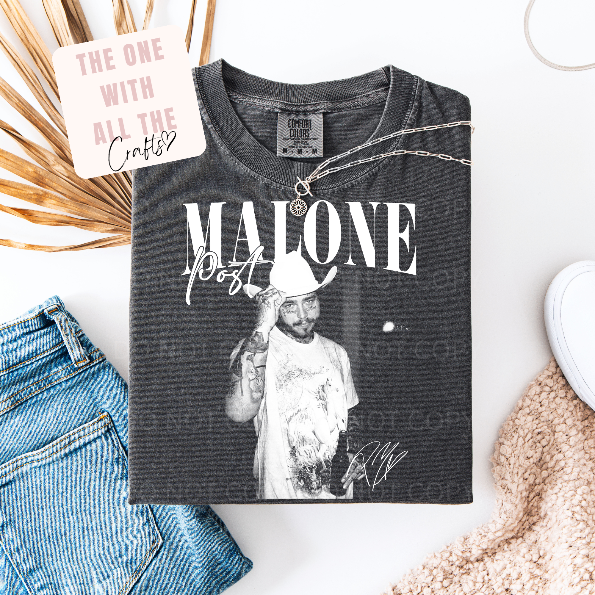 Black t-shirt with graphic design and text, paired with jeans and a necklace on a white surface.