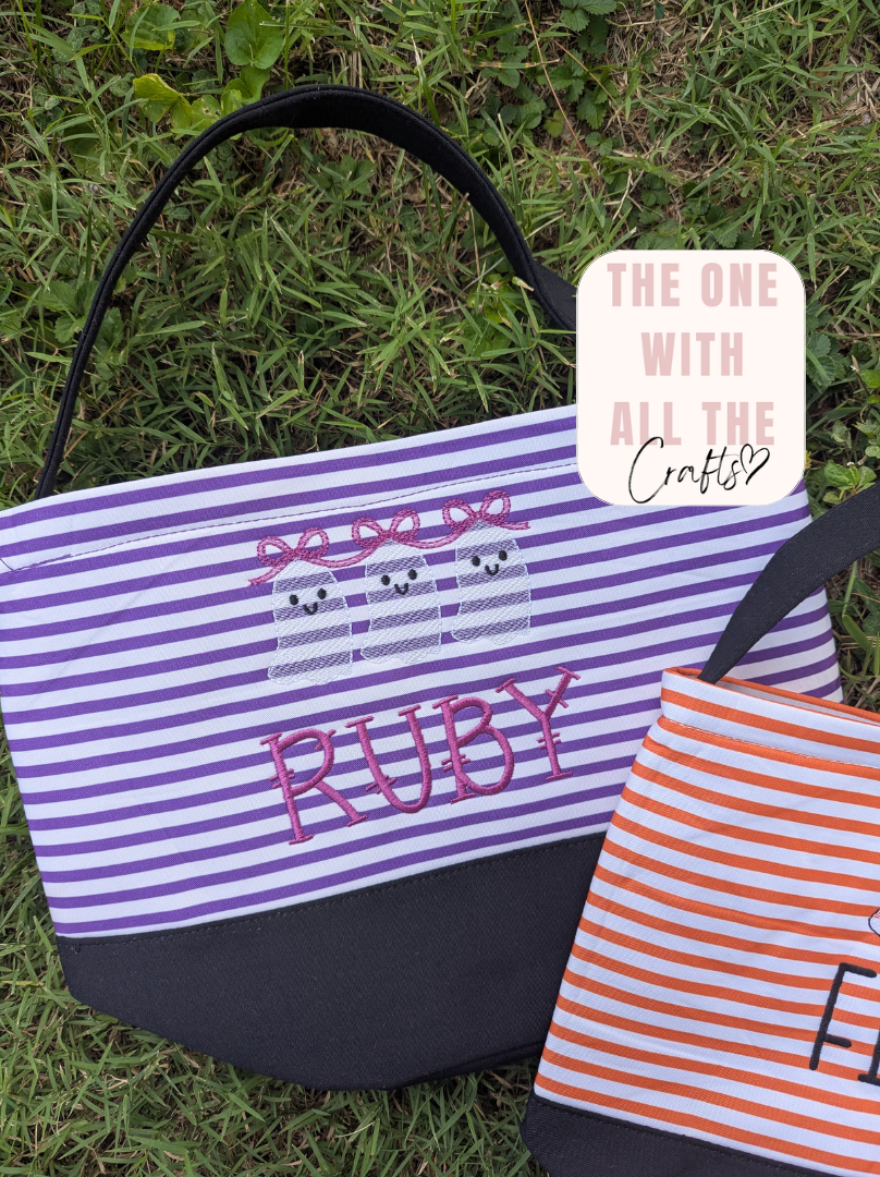 Two personalized canvas tote bags displayed on green grass. The larger bag in the foreground features purple and white horizontal stripes, a black bottom panel, and pink embroidered text reading 'RUBY' along with a ghost design.