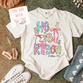 White t-shirt with colorful text design on a textured surface with accessories.