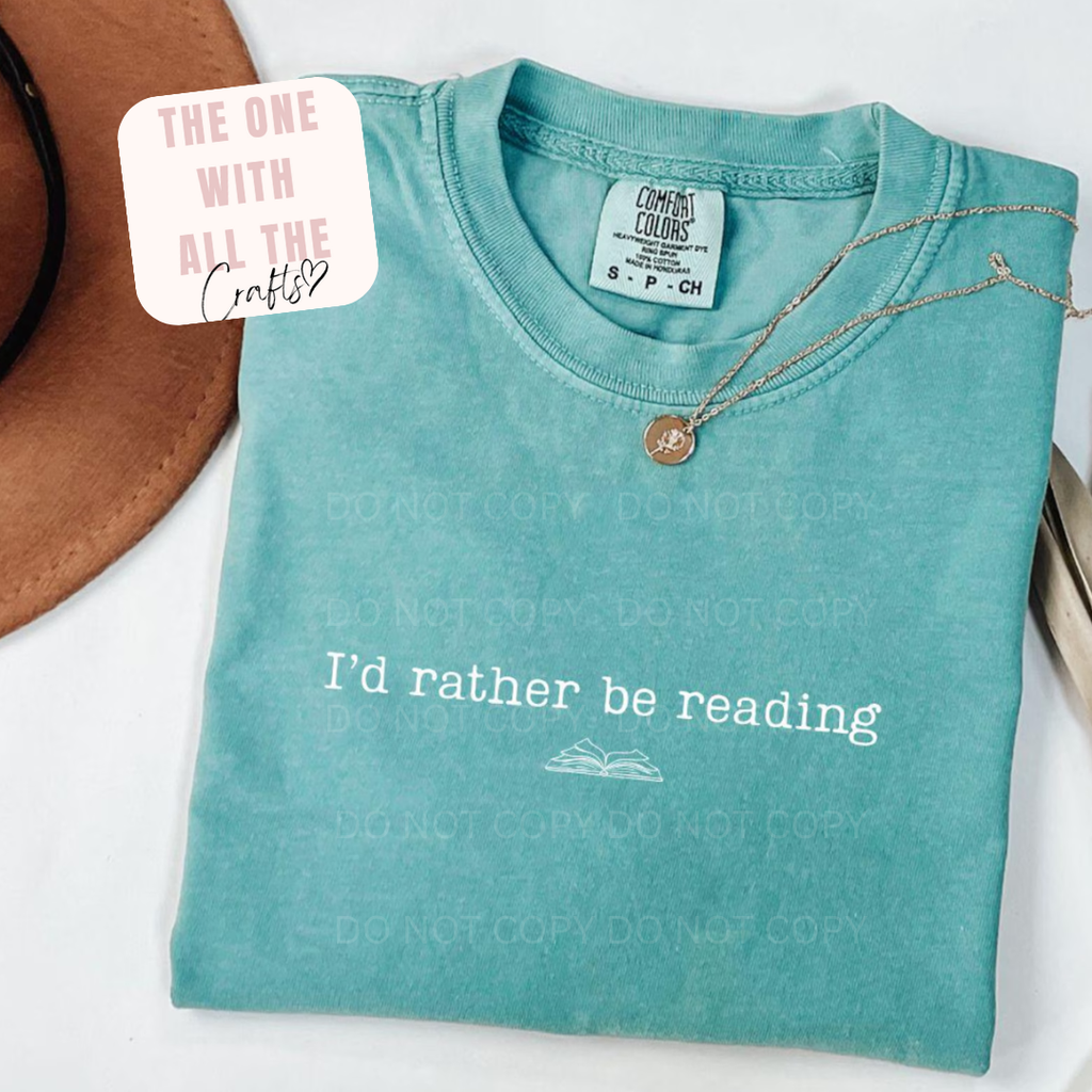 Folded teal t-shirt with 'I'd rather be reading' text and a necklace on a white background