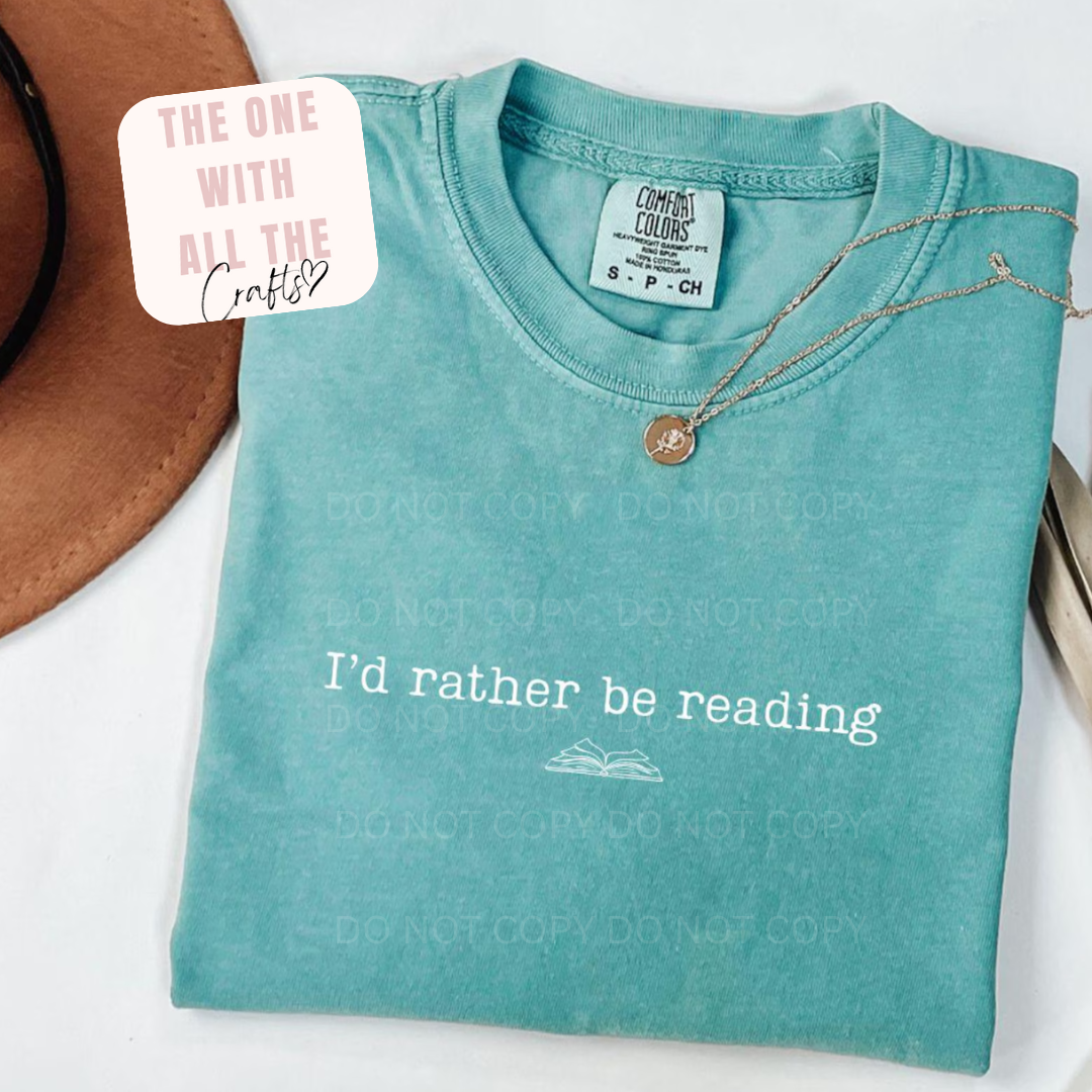 Folded teal t-shirt with 'I'd rather be reading' text and a necklace on a white background