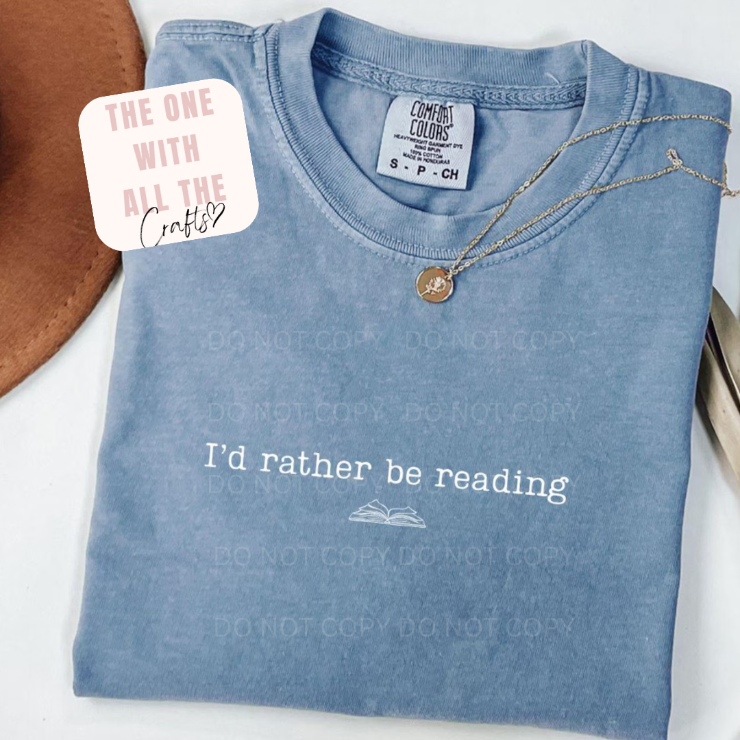 Folded blue t-shirt with 'I'd rather be reading' text and a necklace on a white surface.