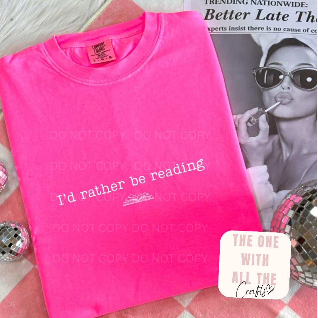 Pink t-shirt with 'I'd rather be reading' text on a surface with a magazine in the background.
