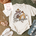 White t-shirt with 'He is risen' faux stitch design with rhinestones featuring Jesus, a cross, and a heart, surrounded by accessories on a textured surface.
