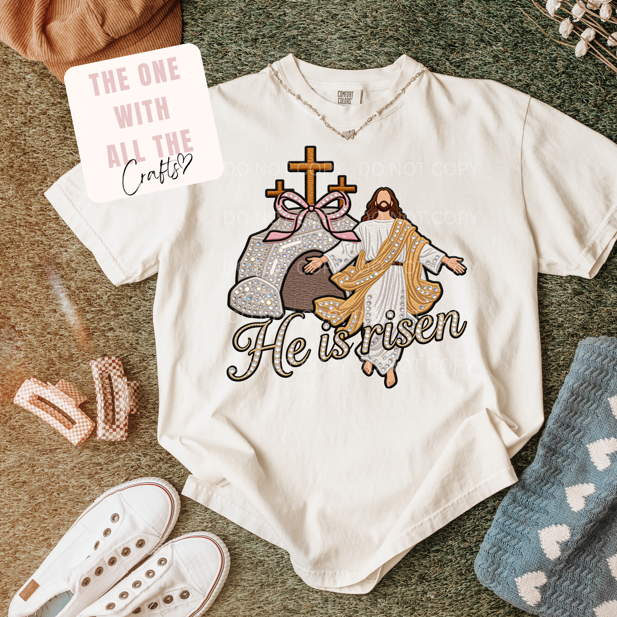 White t-shirt with 'He is risen' faux stitch design with rhinestones featuring Jesus, a cross, and a heart, surrounded by accessories on a textured surface.
