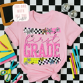Pink t-shirt with 'first grade' design on a checkered background with craft supplies.