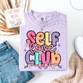 Purple t-shirt with 'Self Love Club' text on a white surface with accessories.