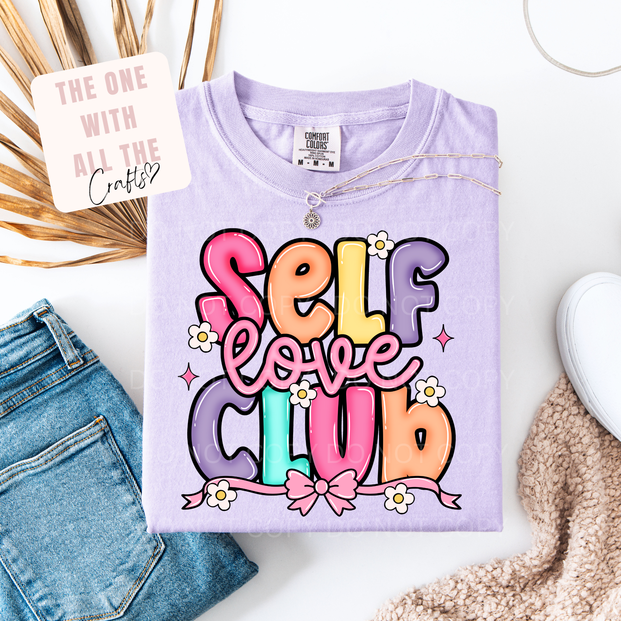 Purple t-shirt with 'Self Love Club' text on a white surface with accessories.