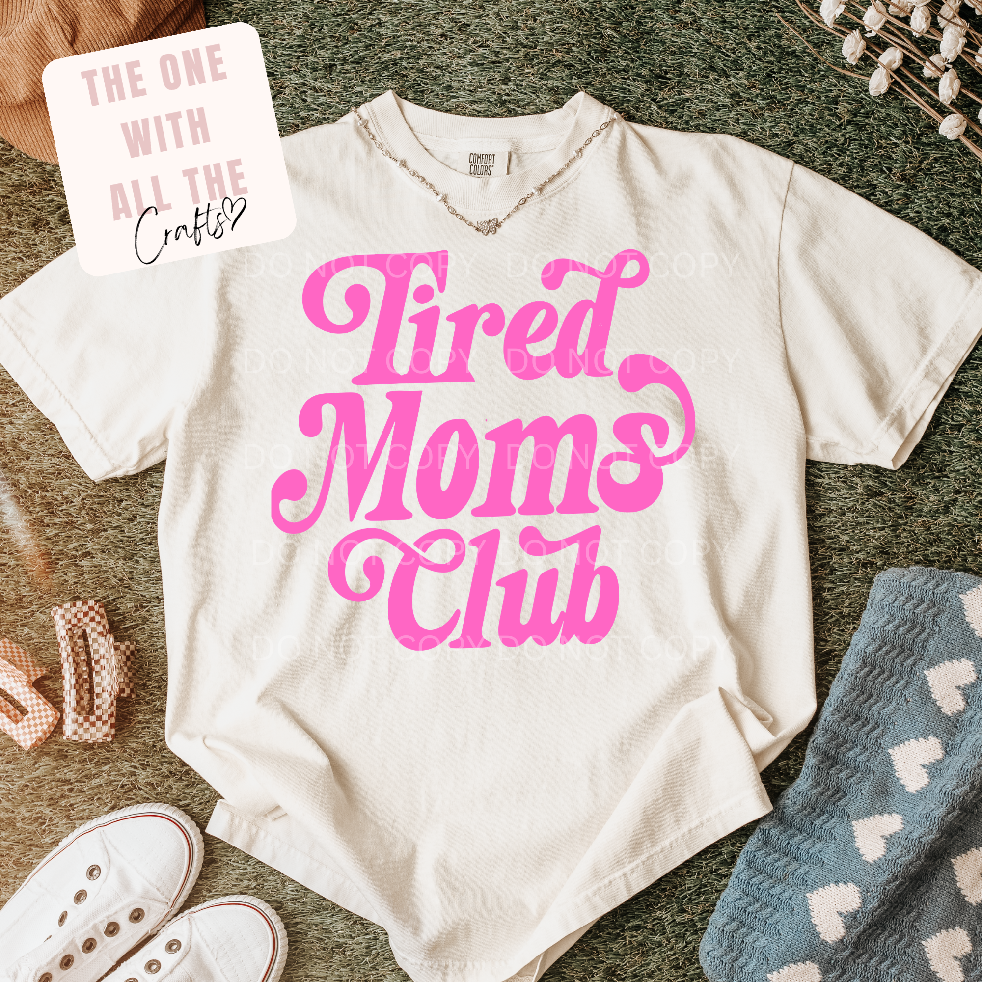 White t-shirt with pink 'Tired Moms Club' text on a textured surface with accessories.