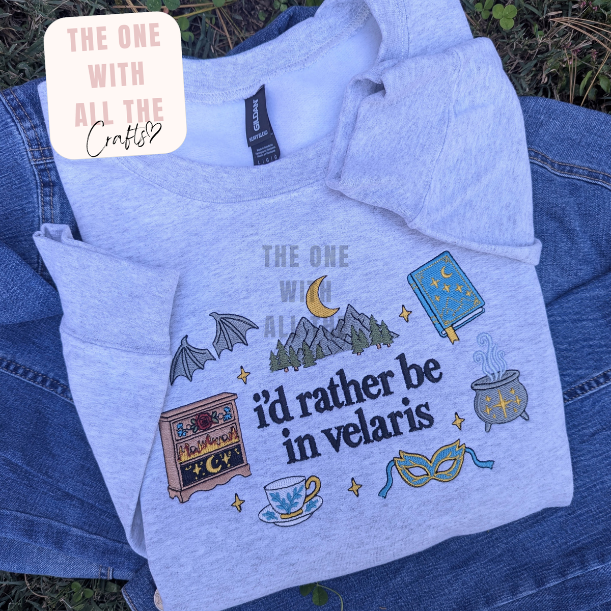 A light heather gray graphic T-shirt laid flat on blue denim, with a decorative printed design including a moon, mountains, bat wings, book, cauldron, mask, teacup, and the phrase “The One With All The i’d Rather Be in Velaris.” A cream badge in the top left reads “The One With All The Crafty?”.