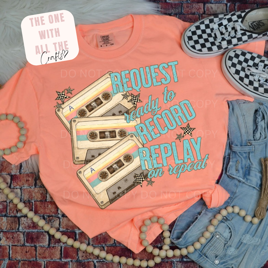 Peach t-shirt with vintage cassette tape design and text on a brick surface with accessories.
