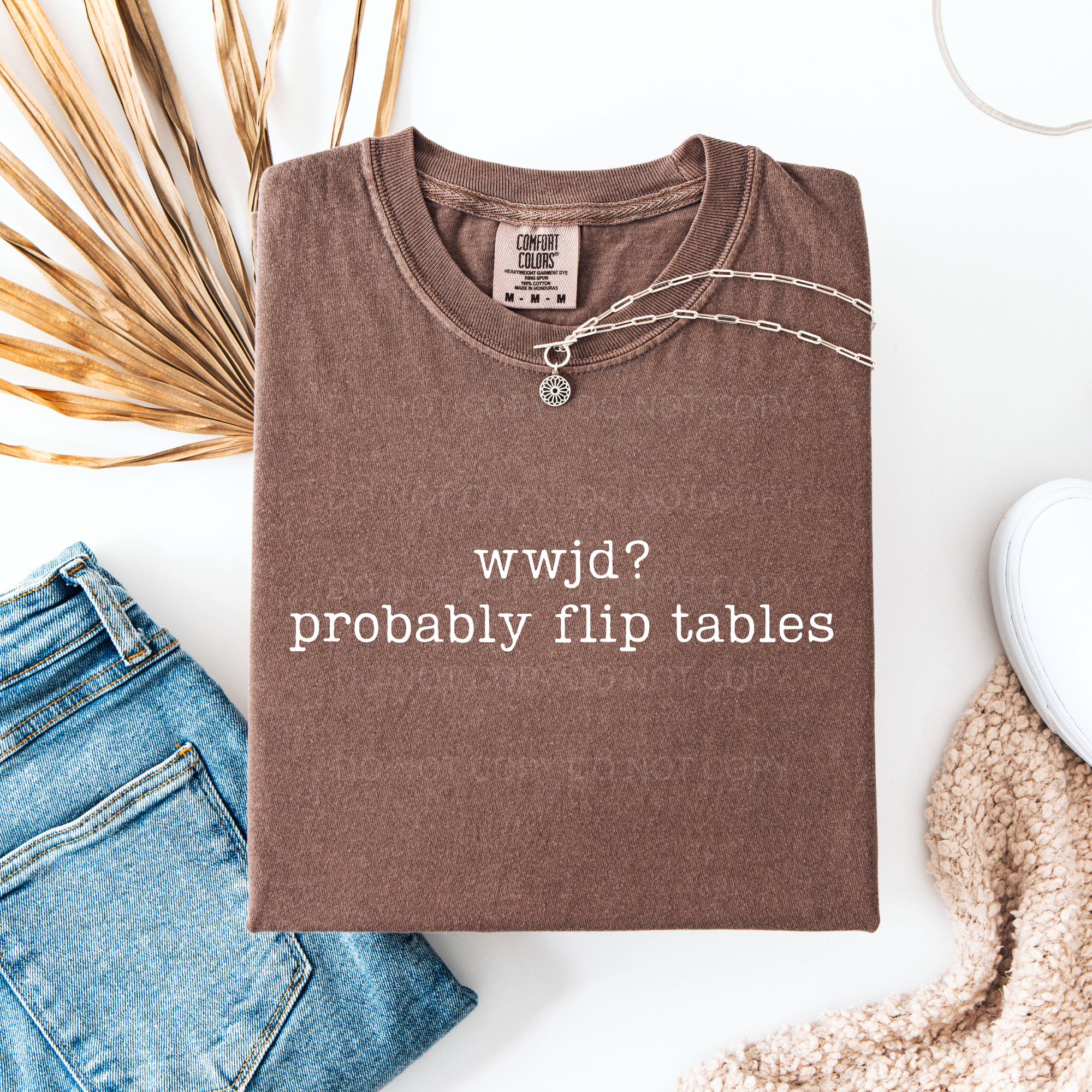 Folded brown t-shirt with text 'wwjd? probably flip tables' on a white surface with jeans and decorative elements.