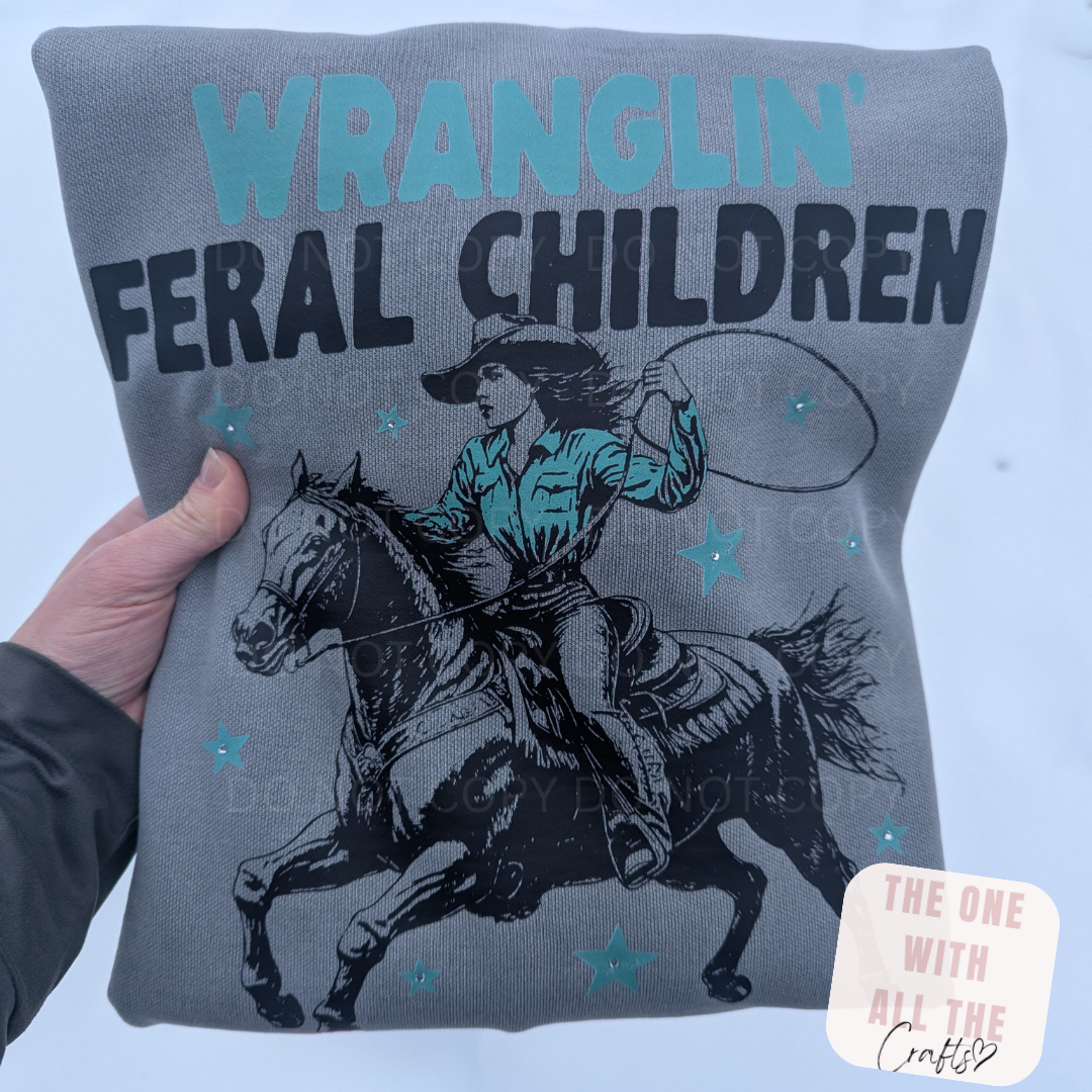 Gray sweatshirt with a graphic of a person riding a horse and text 'WRANGLIN' FERAL CHILDREN' held by a hand against a light blue background.