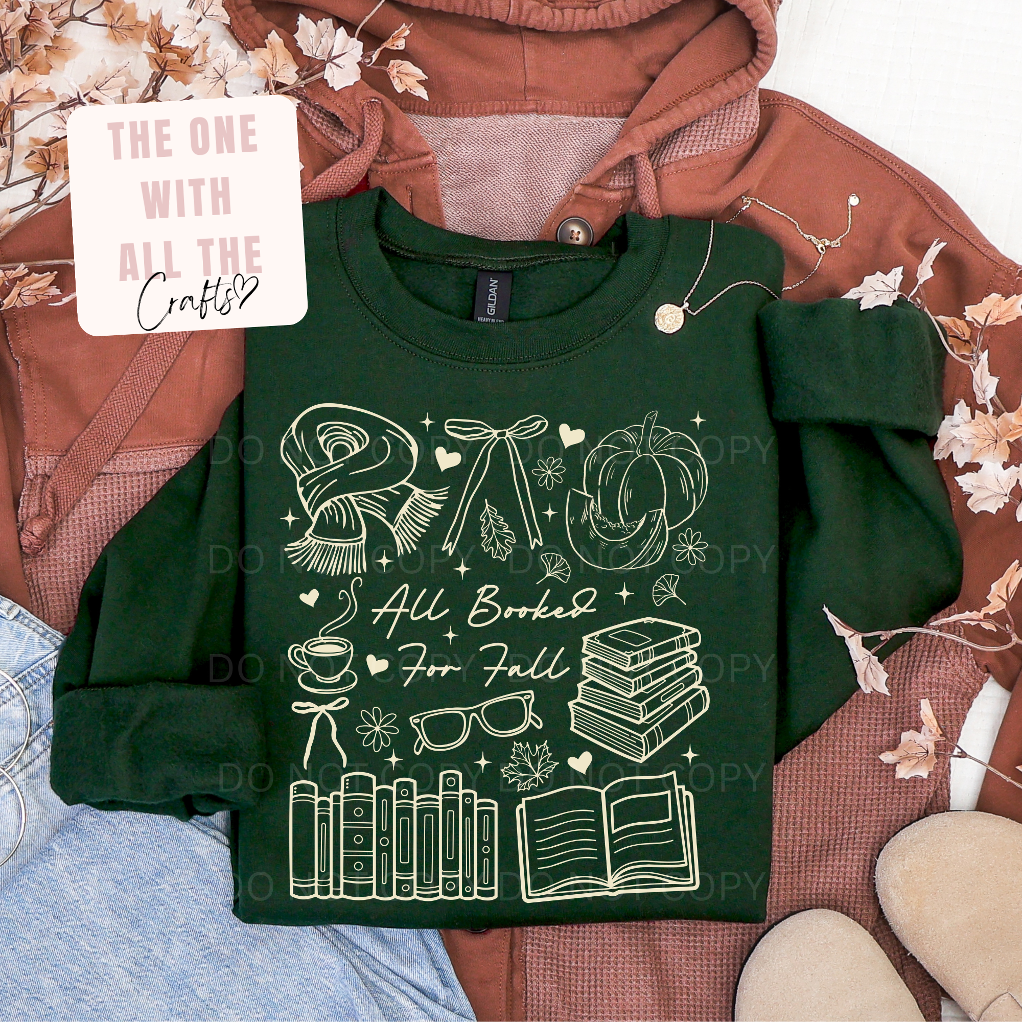 A dark green crewneck sweatshirt with a white autumn-themed graphic reading 'All Baked For Fall' surrounded by illustrations of a scarf, pumpkin, books, and glasses, styled in a flat lay with a rust-colored hoodie, denim, and a decorative card.