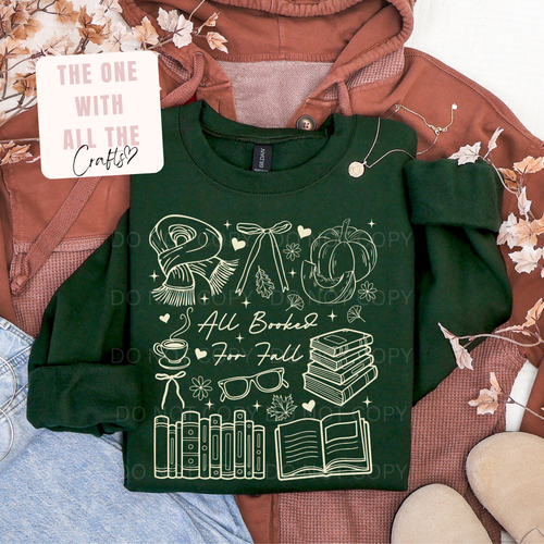 A dark green crewneck sweatshirt with a white autumn-themed graphic reading 'All Baked For Fall' surrounded by illustrations of a scarf, pumpkin, books, and glasses, styled in a flat lay with a rust-colored hoodie, denim, and a decorative card.