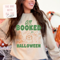 A woman wearing a cream-colored crewneck sweatshirt with a green and orange graphic that reads 'all BOOKED for HALLOWEEN' with illustrations of a ghost reading, a witch hat, candles, and a pumpkin. A white sticker in the top left corner reads 'THE ONE WITH ALL THE Crafts?'.