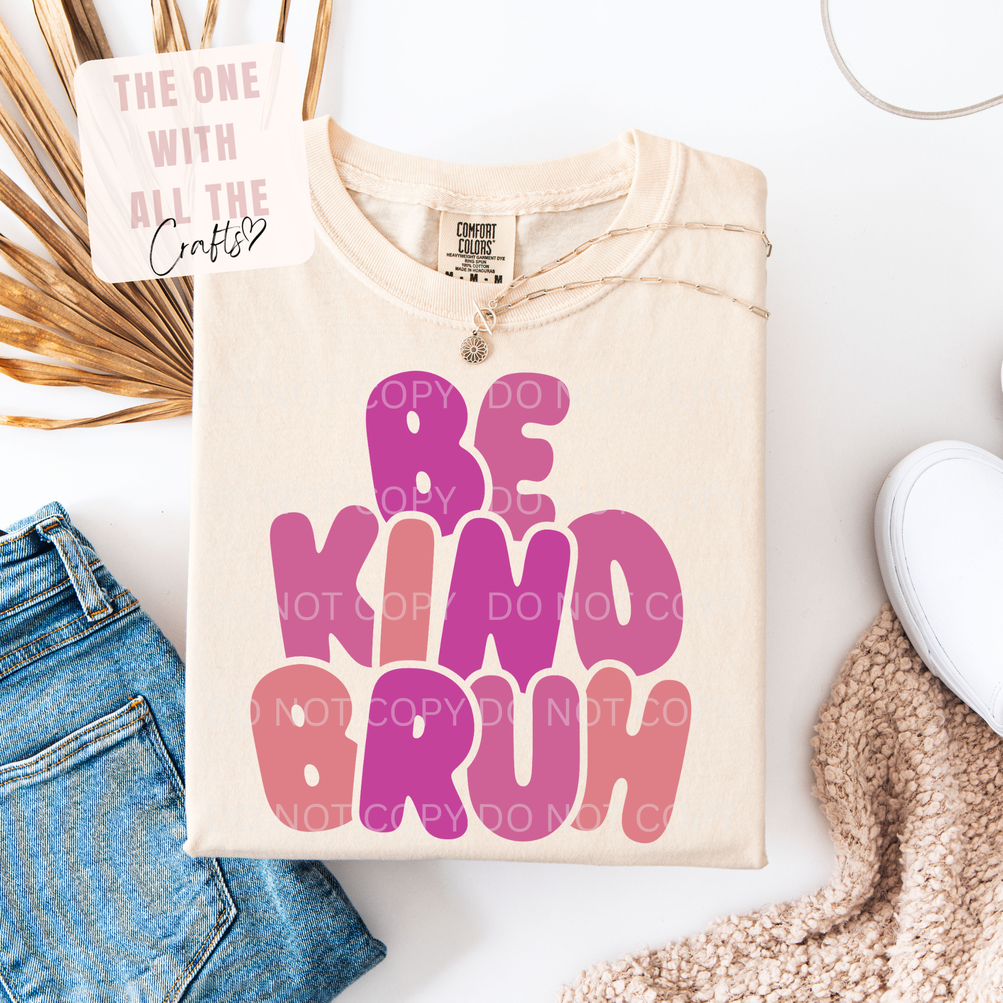 A folded cream-colored t-shirt with 'BE KIND BRUH' printed in bold pink and purple bubble letters, styled in a flat lay with blue jeans, a white sneaker, a knit blanket, and a decorative card.