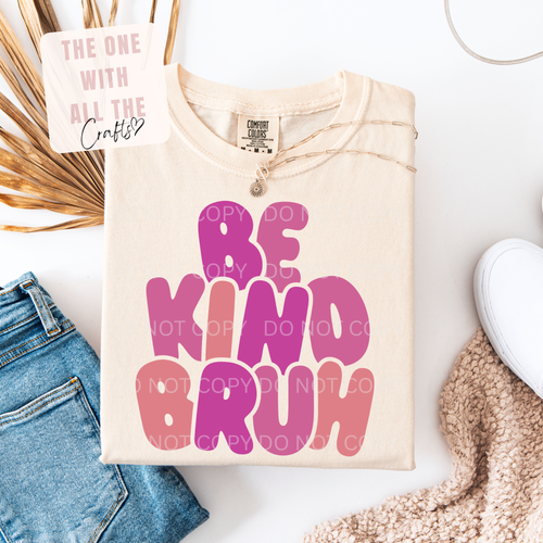 A folded cream-colored t-shirt with 'BE KIND BRUH' printed in bold pink and purple bubble letters, styled in a flat lay with blue jeans, a white sneaker, a knit blanket, and a decorative card.